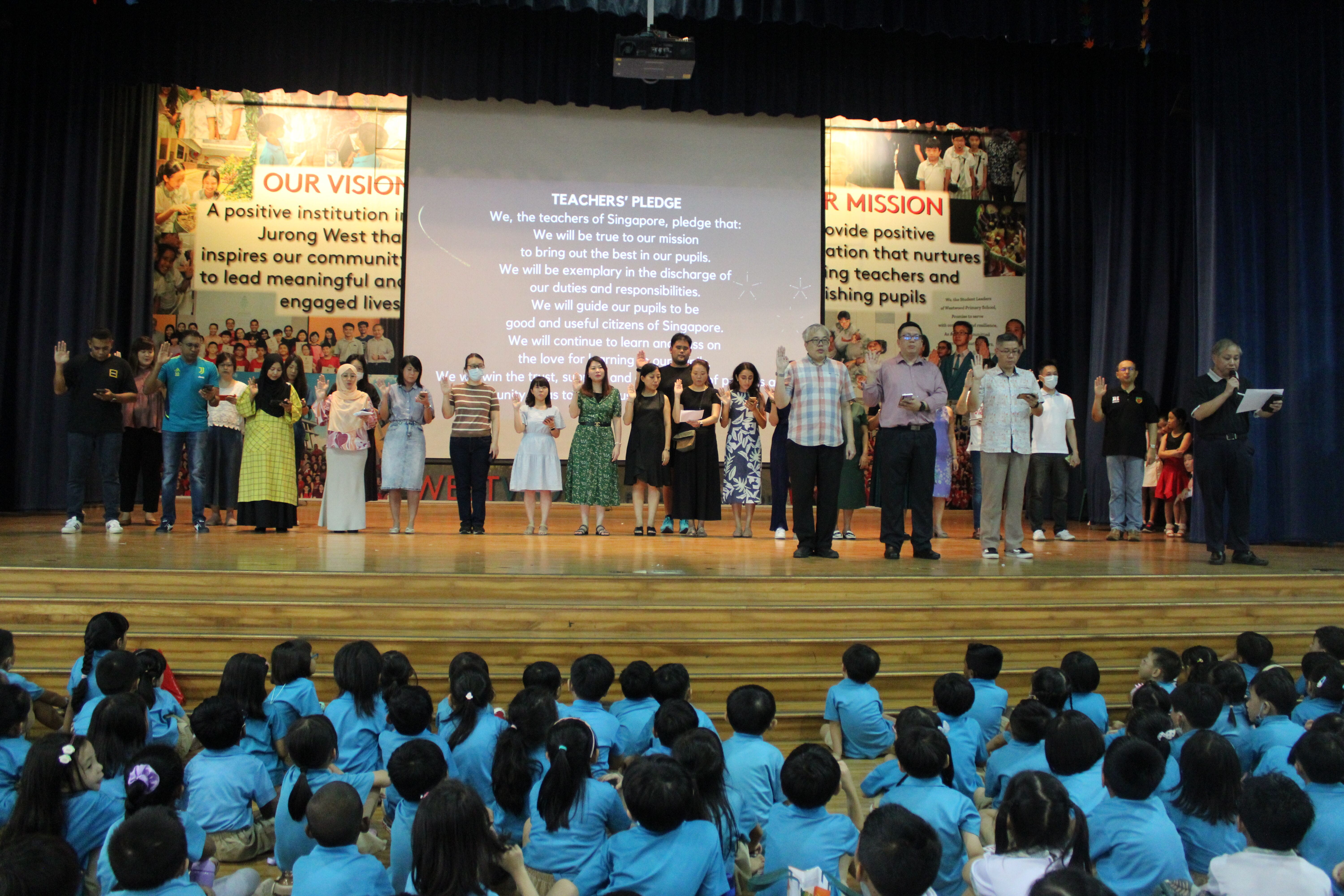 Teachers stand on a stage reciting the Teachers' Pledge before students in blue uniforms seated on the floor.